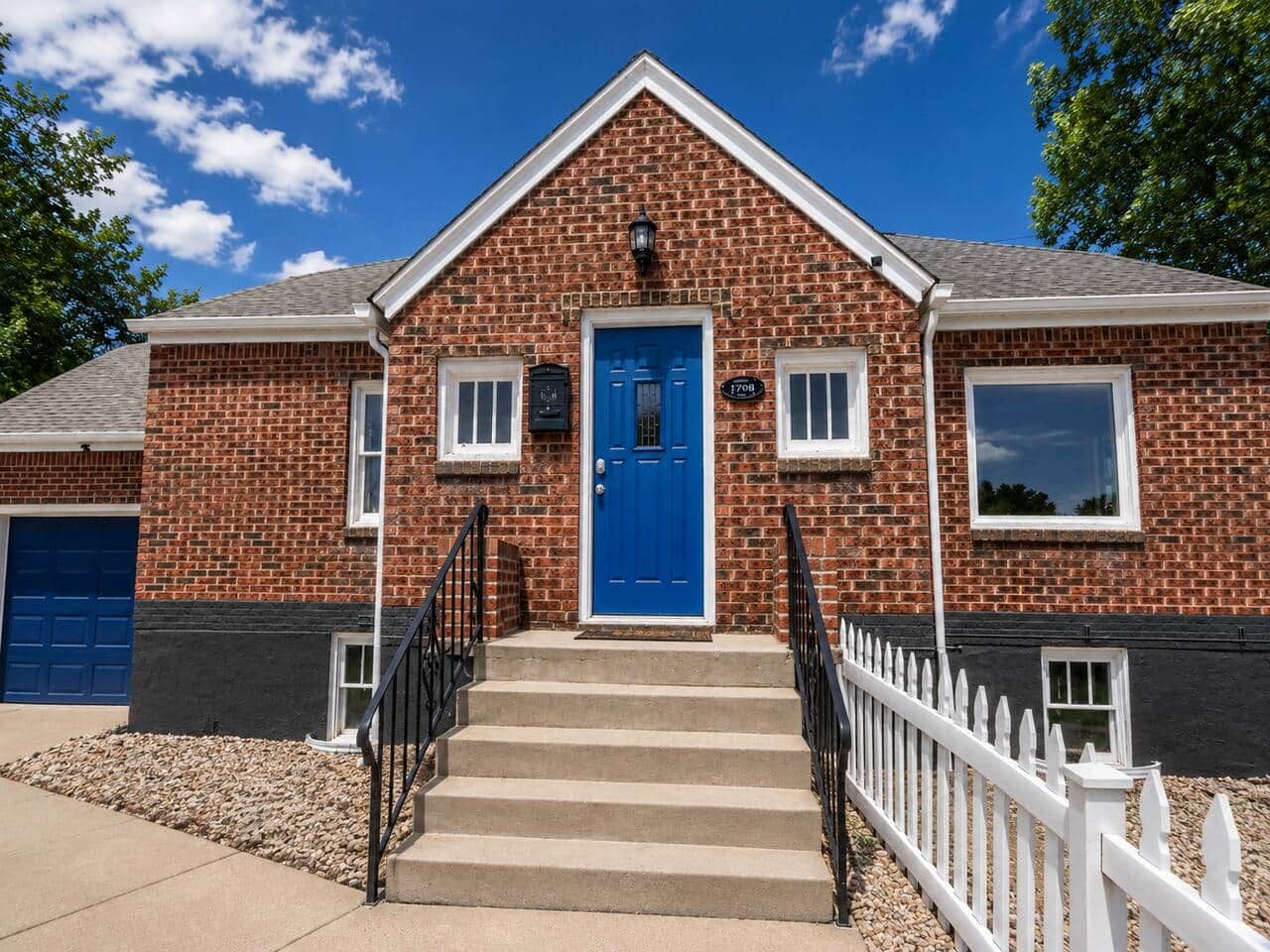 Une maison à deux étages, construite en briques, avec une porte d’entrée bleue, des balustrades blanches et un petit porche.