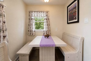Um quarto pequeno com um banco branco, uma mesa com toalha roxa e uma janela com cortinas.