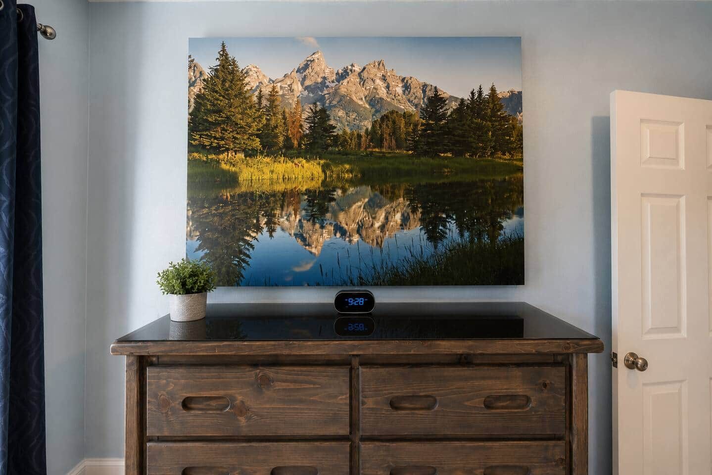 A wall-mounted television displaying a mountain landscape, a digital clock, and a small potted plant on a wooden dresser.