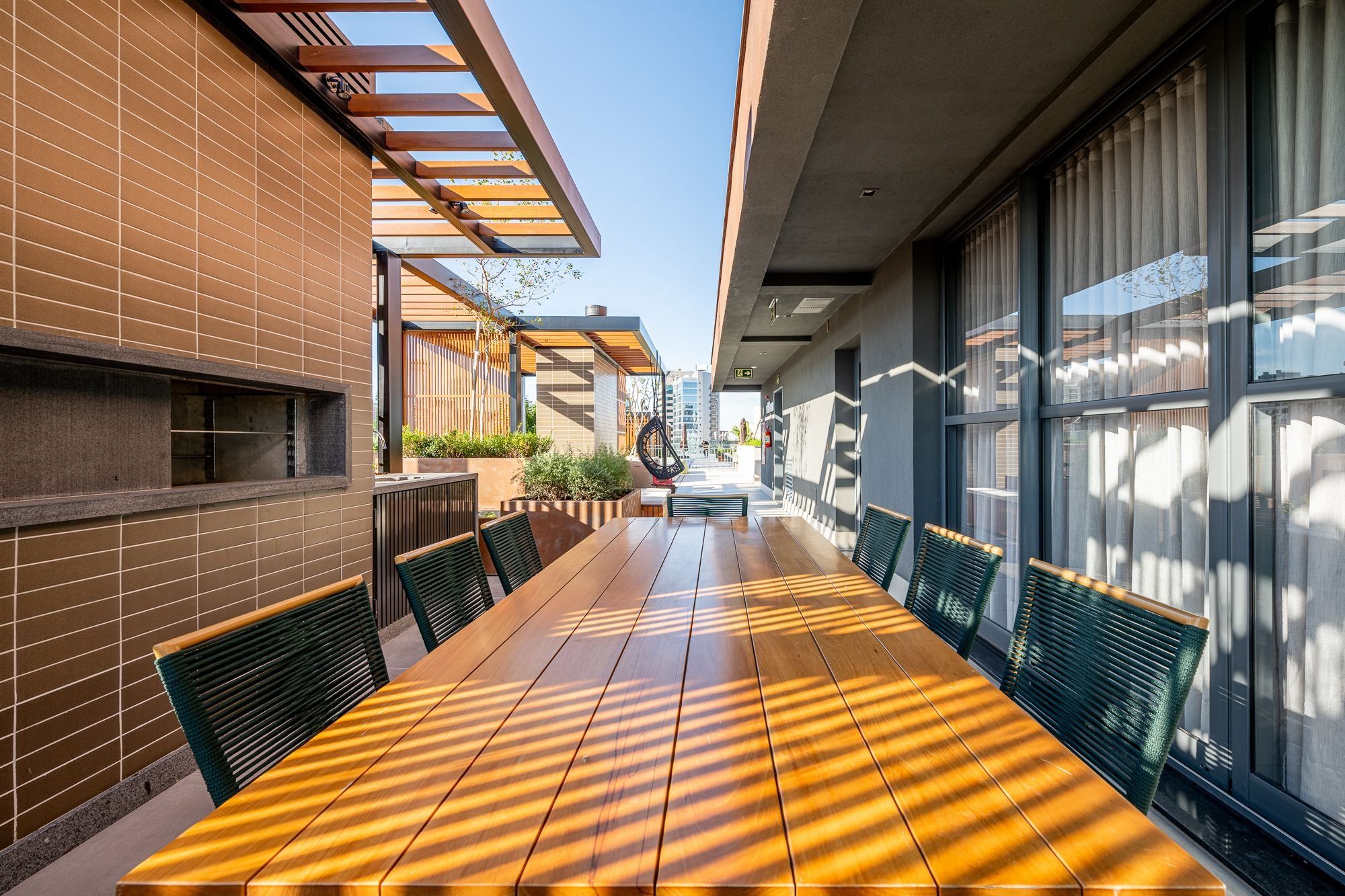 Un área de comedor al aire libre con una larga mesa de madera y sillas, una pared de ladrillo y ventanas grandes.