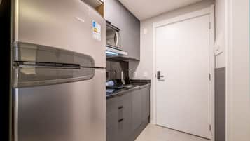 Cocina compacta con refrigerador de acero inoxidable, microondas y horno integrado. Se observa una puerta blanca a la derecha y una encimera con lavamanos.