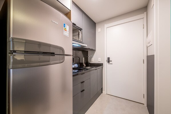 A compact kitchen with a stainless steel refrigerator, a microwave, and a built-in oven. There is a white door to the right and a countertop with a sink.