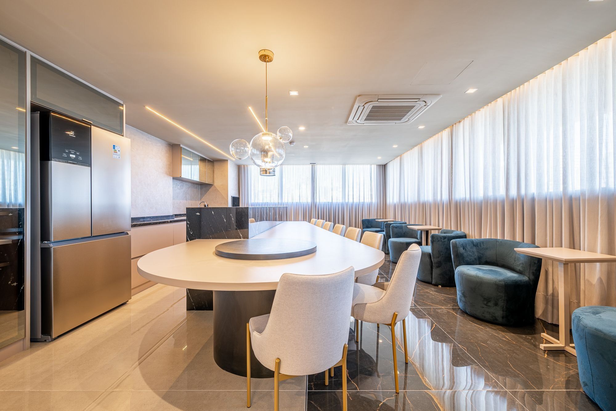 A modern hotel dining area with a central round table, seating, and a kitchen in the background.