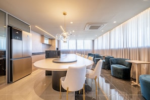 Un comedor moderno de hotel con una mesa redonda central, asientos y una cocina al fondo.
