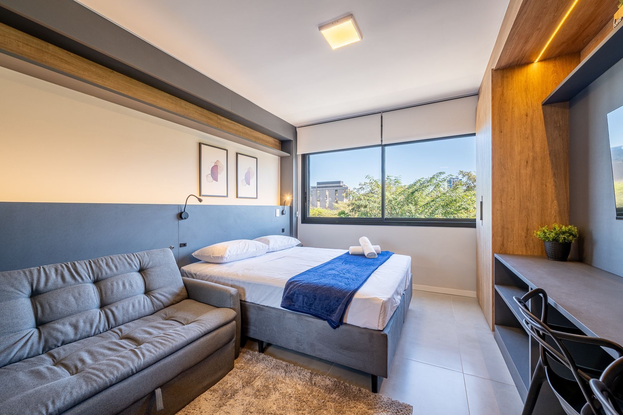 A modern hotel room with a bed, a sofa, a desk, and a large window offering a view of the outdoors.