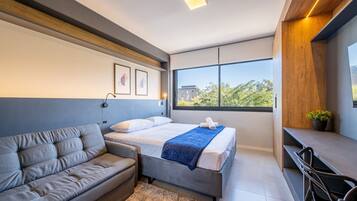 A modern hotel room with a bed, a sofa, a desk, and a large window offering a view of the outdoors.