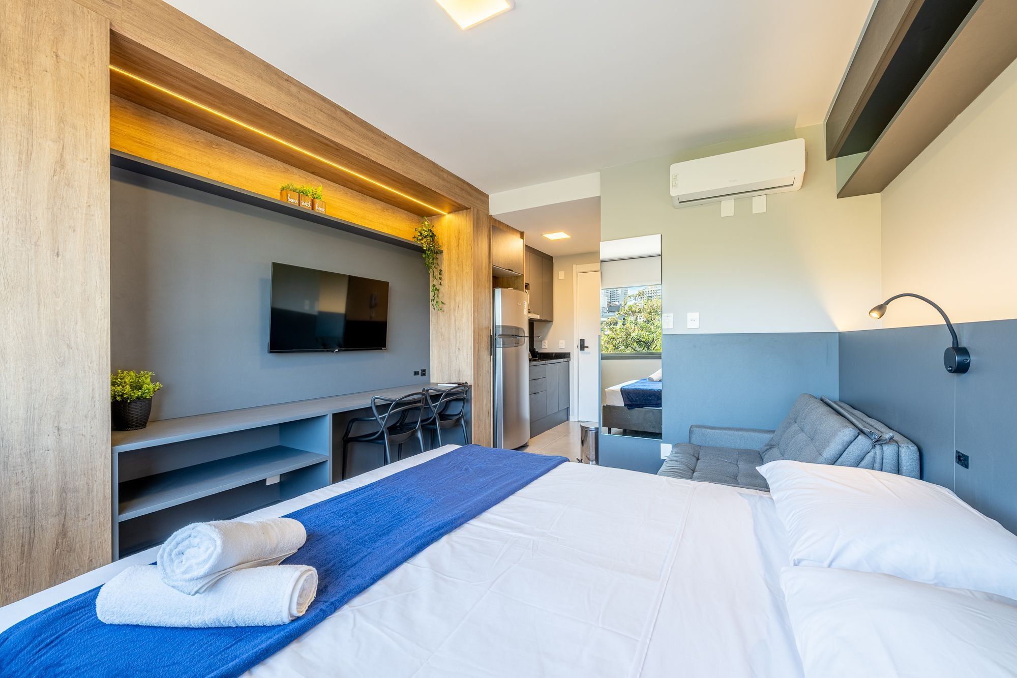 Quarto de hotel moderno com cama, televisão, escrivaninha e cozinha compacta.
