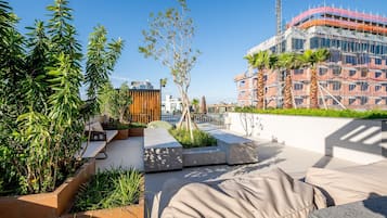 Una terraza en la azotea con mobiliario exterior, vegetación y vistas a un edificio en construcción.