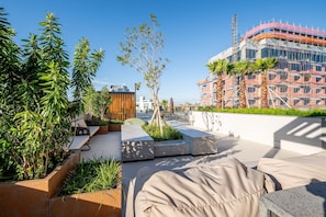 Una terraza en la azotea con mobiliario exterior, vegetación y vistas a un edificio en construcción.