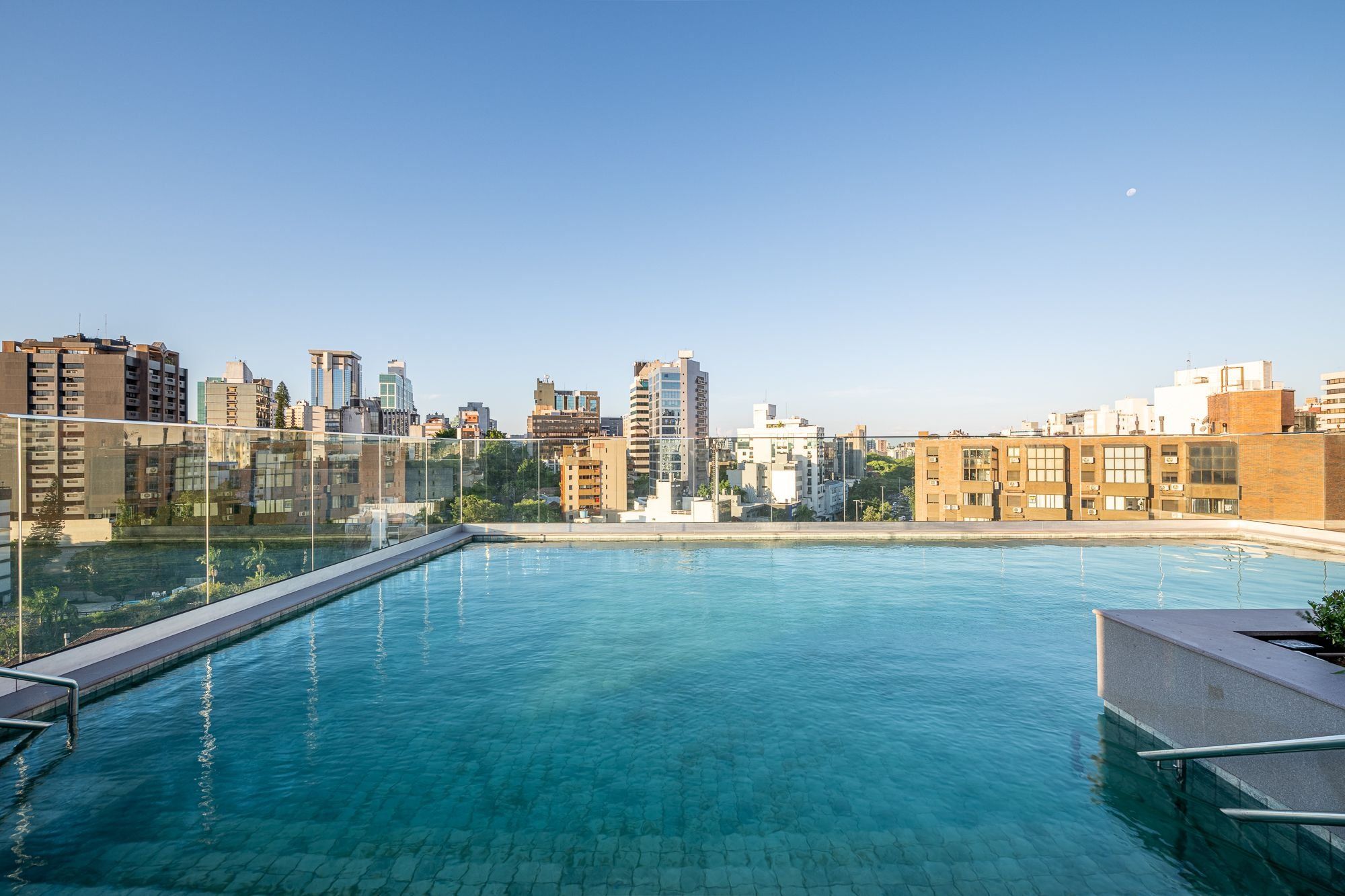 Piscina ao ar livre com água azul cristalina, cercada por uma barreira de vidro e com vista para um horizonte urbano repleto de edifícios.