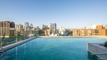 Piscina ao ar livre com água azul cristalina, cercada por uma barreira de vidro e com vista para um horizonte urbano repleto de edifícios.