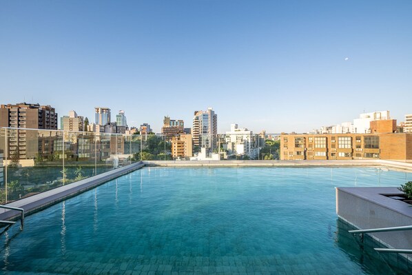 Piscina exterior con agua azul cristalina, rodeada por una barrera de vidrio y con vistas a un paisaje urbano con varios edificios.