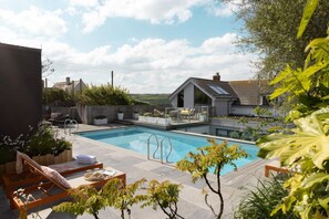 A swimming pool with lounge chairs and a deck area, surrounded by greenery and a view of a hill. - 6 Bed in Mawgan Porth (oc-b584421) (Mawgan Porth)