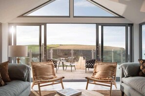 A spacious room with a high ceiling, large windows, and a view of the countryside. There are two wicker chairs and a sofa. - 6 Bed in Mawgan Porth (oc-b584421) (Mawgan Porth)