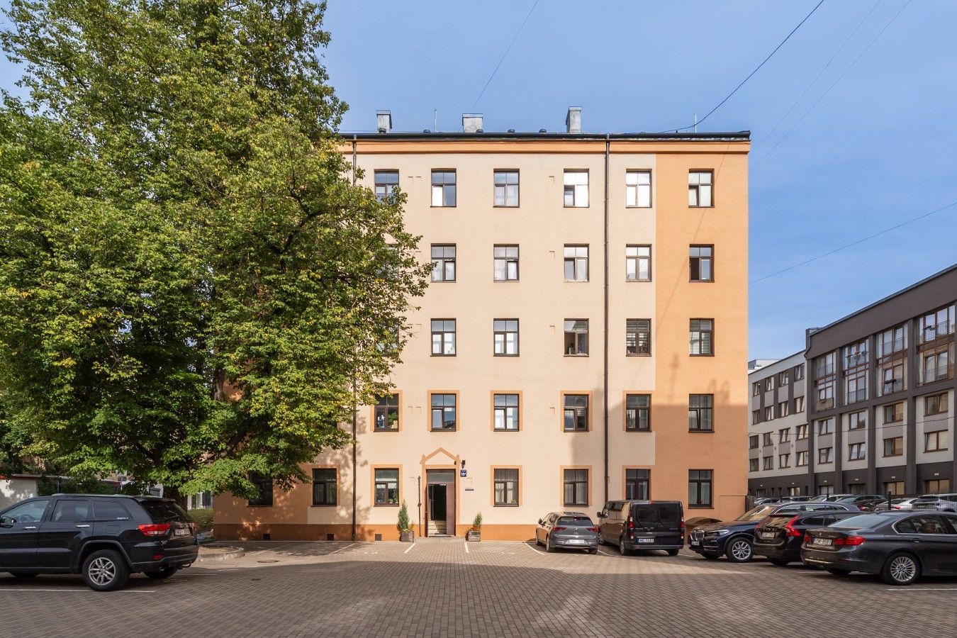 Un immeuble à plusieurs étages doté d’une façade plate, d’un parking à l’avant et d’un grand arbre sur le côté gauche.