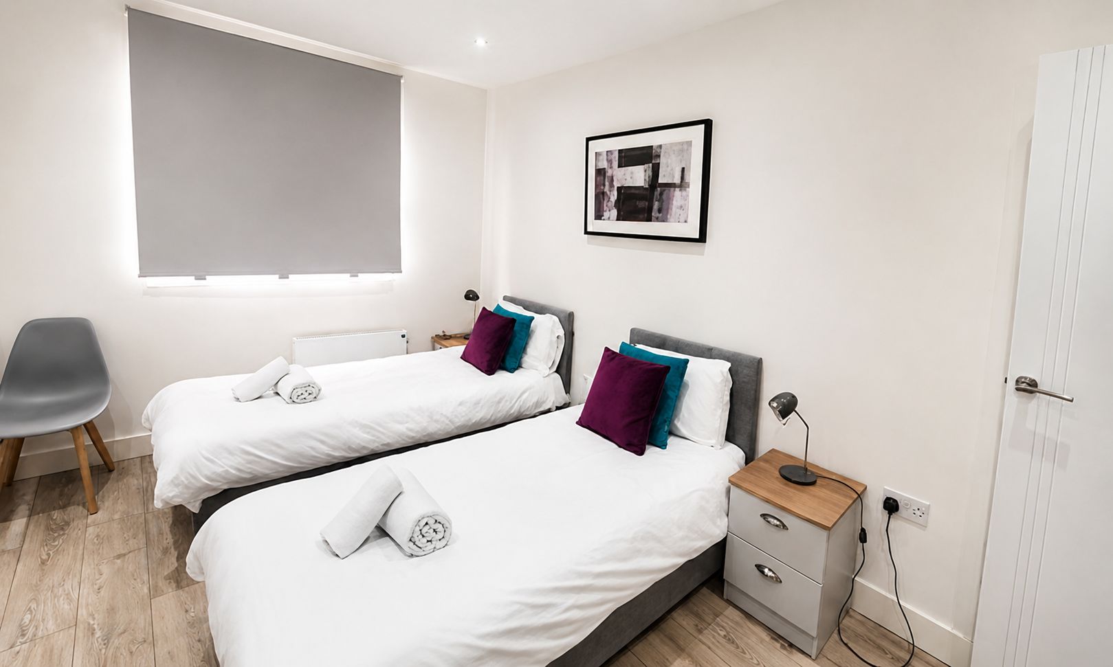 A hotel room with two beds, a desk, a chair, a window with a blind, a picture on the wall, and a door.