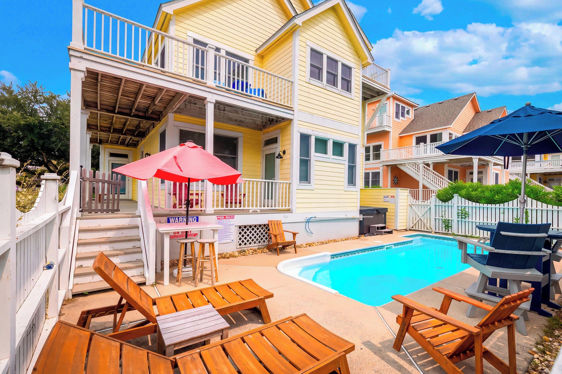 A poolside area with wooden chairs and a table, a small pool, and a building with a balcony.