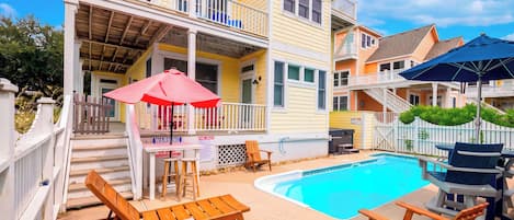 A poolside area with wooden chairs and a table, a small pool, and a building with a balcony.