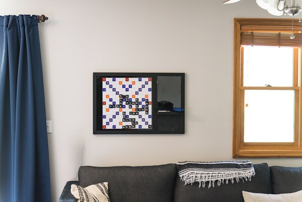 A living room with a framed Scrabble board on the wall, a window with blinds, and a dark-colored sofa with a throw blanket.