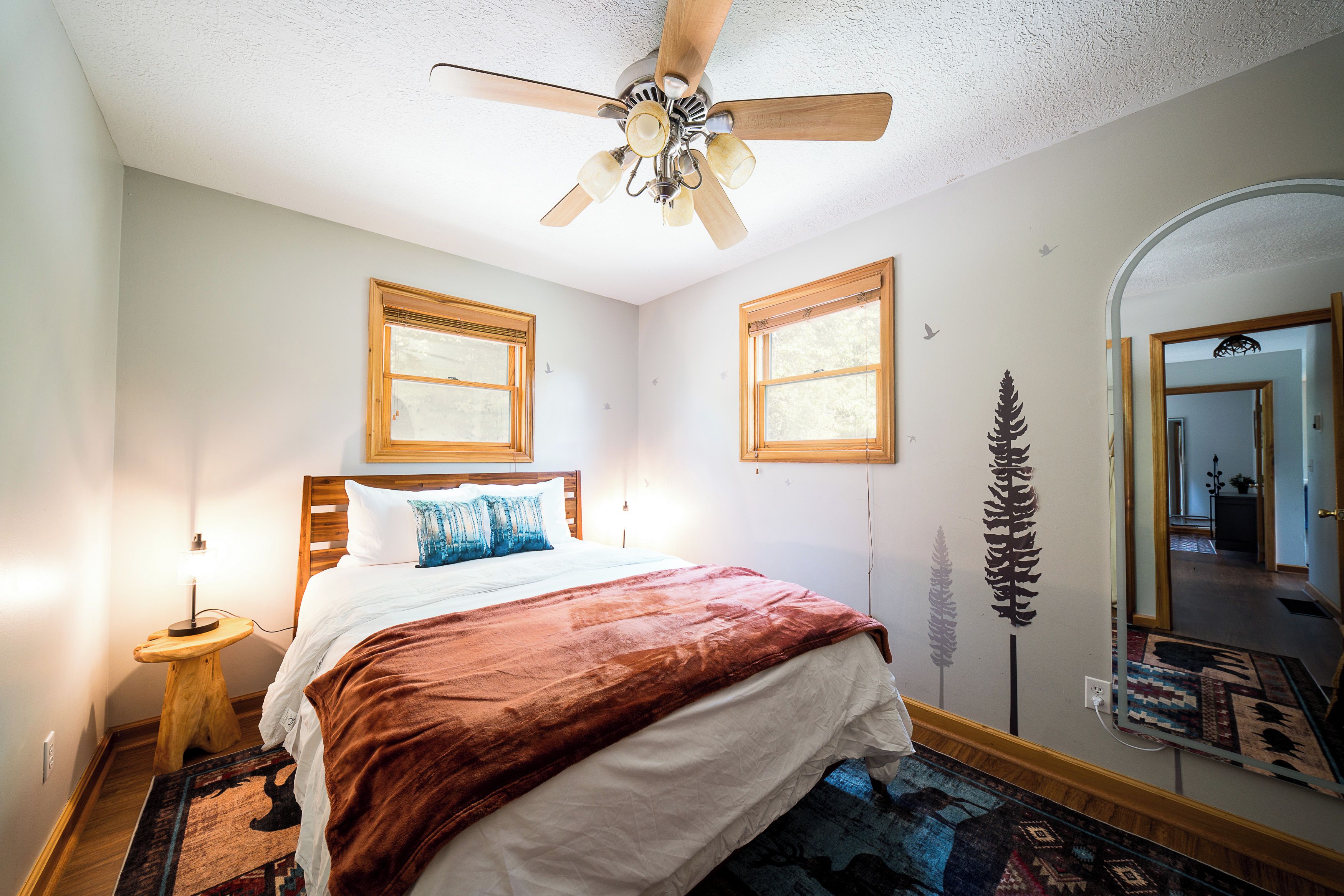 Une chambre à coucher avec un lit, un ventilateur de plafond, deux fenêtres, un miroir et un tapis.