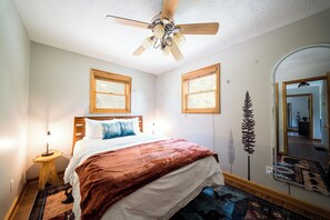 A bedroom with a bed, a ceiling fan, two windows, a mirror, and a rug.