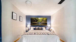 Quarto com cama, duas luminárias de cabeceira, televisão fixada na parede e quadros emoldurados.
