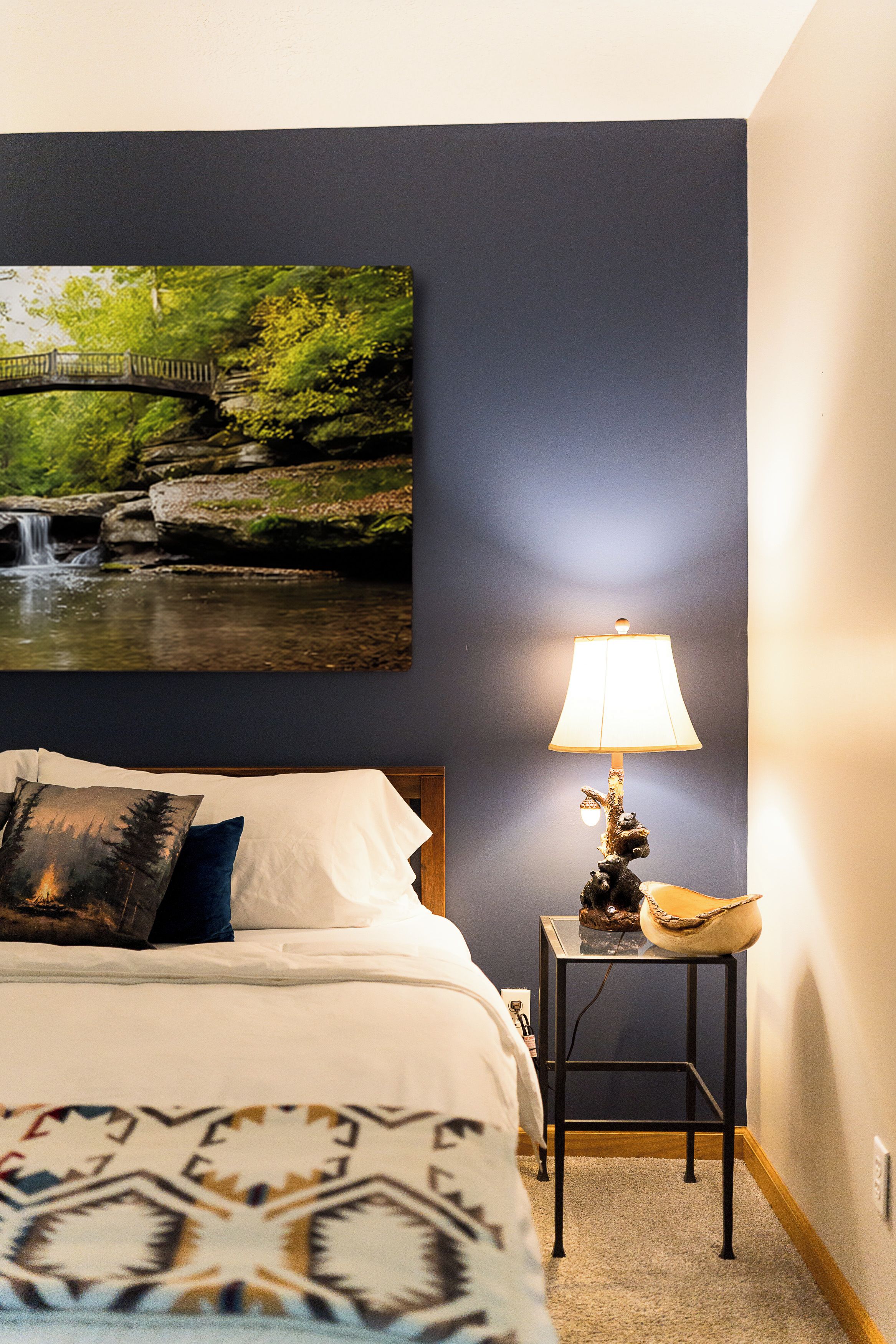 Une chambre à coucher avec un lit, une table de chevet avec une lampe, un tableau représentant un pont au-dessus d’une cascade et un plaid à motifs.