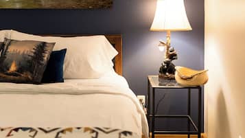 Une chambre à coucher avec un lit, une table de chevet avec une lampe, un tableau représentant un pont au-dessus d’une cascade et un plaid à motifs.