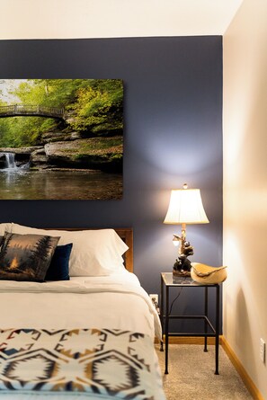A bedroom with a bed, a nightstand with a lamp, a painting of a bridge over a waterfall, and a patterned throw blanket.