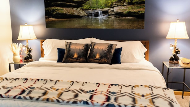 Quarto com cama, duas luminárias de cabeceira, pintura pendurada na parede com tema de cachoeira e colcha estampada.