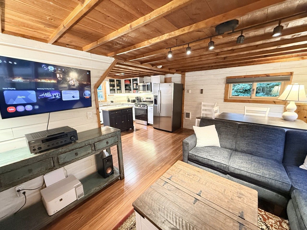 A modern living room with a flat-screen TV, wooden flooring, a gray sofa, and a kitchen in the background.