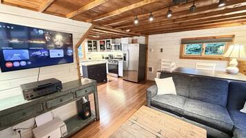 A modern living room with a flat-screen TV, wooden flooring, a gray sofa, and a kitchen in the background.