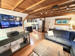 A modern living room with a flat-screen TV, wooden flooring, a gray sofa, and a kitchen in the background.
