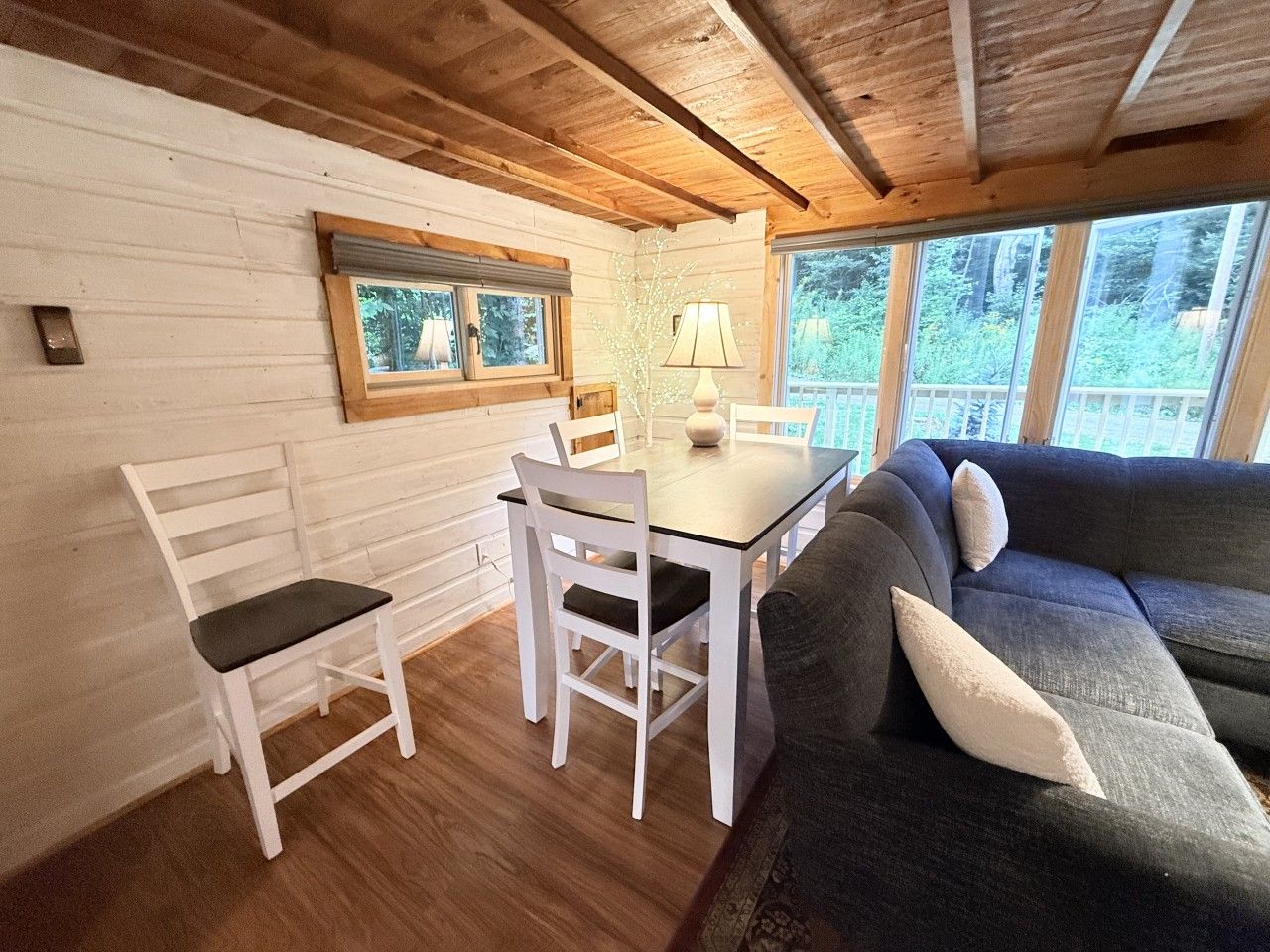 A cozy cabin interior with a dining table, white chairs, a gray sofa, and large windows offering a view of the forest.
