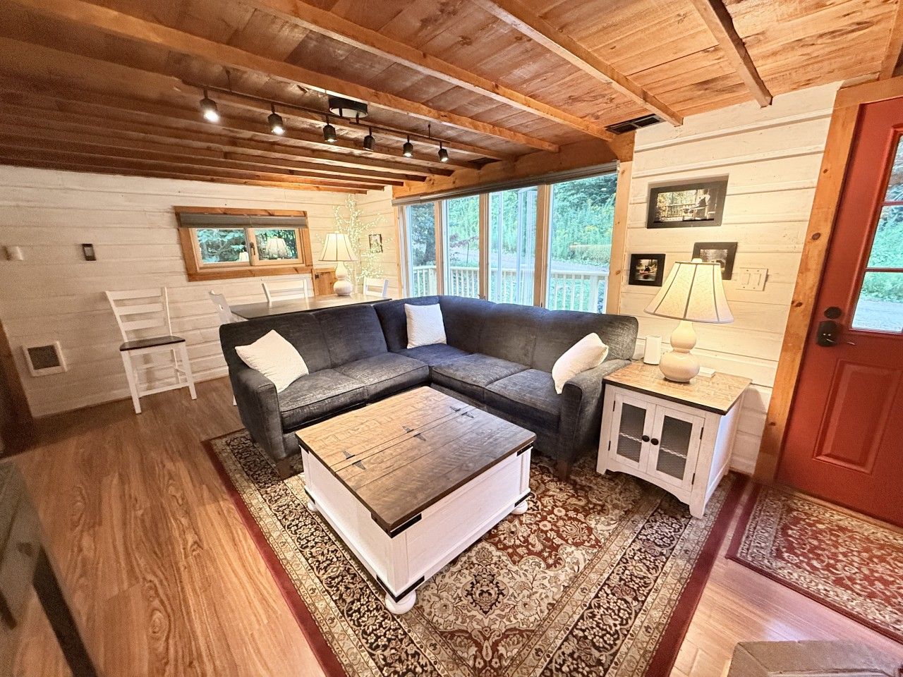 A cozy cabin interior with a sectional sofa, a coffee table, a small cabinet, and a window with a view of the outdoors.