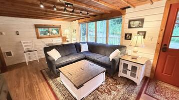 A cozy cabin interior with a sectional sofa, a coffee table, a small cabinet, and a window with a view of the outdoors.
