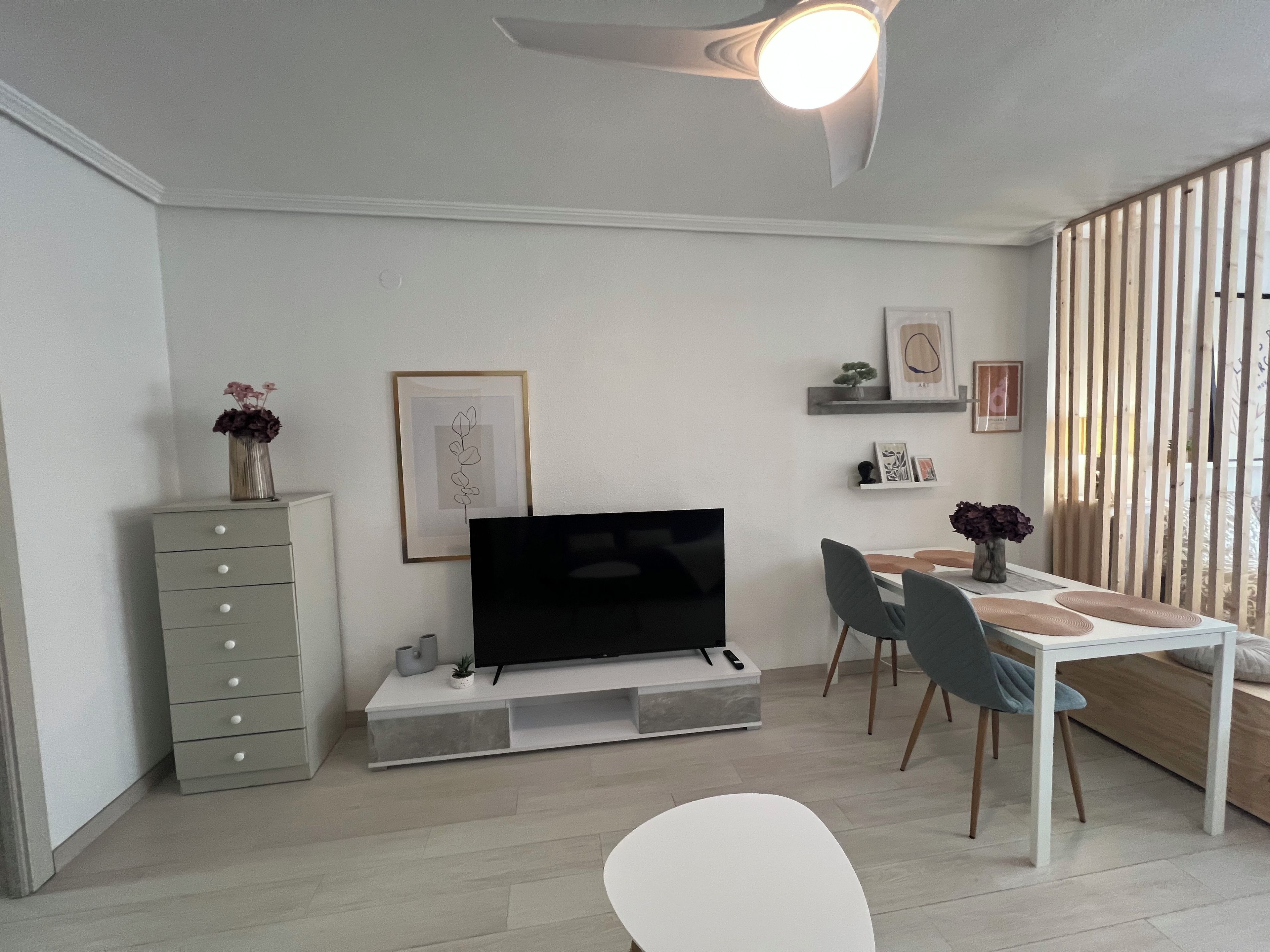A modern living room with a flat-screen TV, a dining table with chairs, a grey sofa, and a ceiling fan.