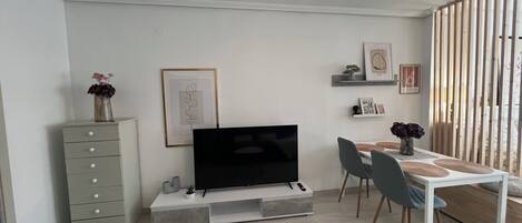 A modern living room with a flat-screen TV, a dining table with chairs, a grey sofa, and a ceiling fan.