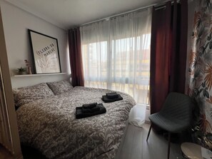 Quarto de hotel com uma cama grande, uma cadeira, janela com cortinas e um quadro na parede.