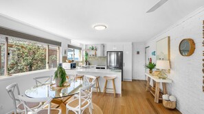A modern kitchen with white cabinetry, a stainless steel refrigerator, and a dining area with a glass table and white chairs. - Plovers Nest (Peregian Beach)