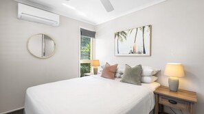 A bedroom with a bed, two bedside lamps, a round mirror, a painting, and an air conditioning unit. - Plovers Nest (Peregian Beach)