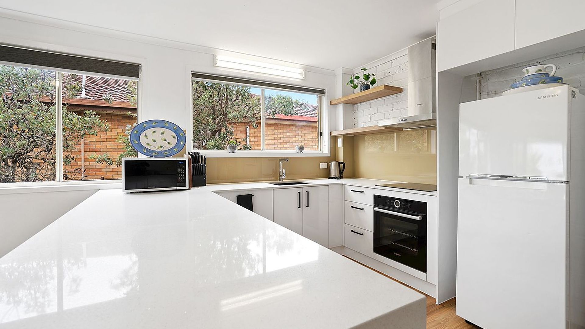 Una cocina moderna con armarios blancos, una isla grande y vistas a una casa de ladrillo a través de las ventanas.