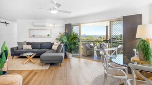 Un salon moderne avec un canapé d’angle gris, une table basse et un coin repas comprenant une table en verre et des chaises blanches.