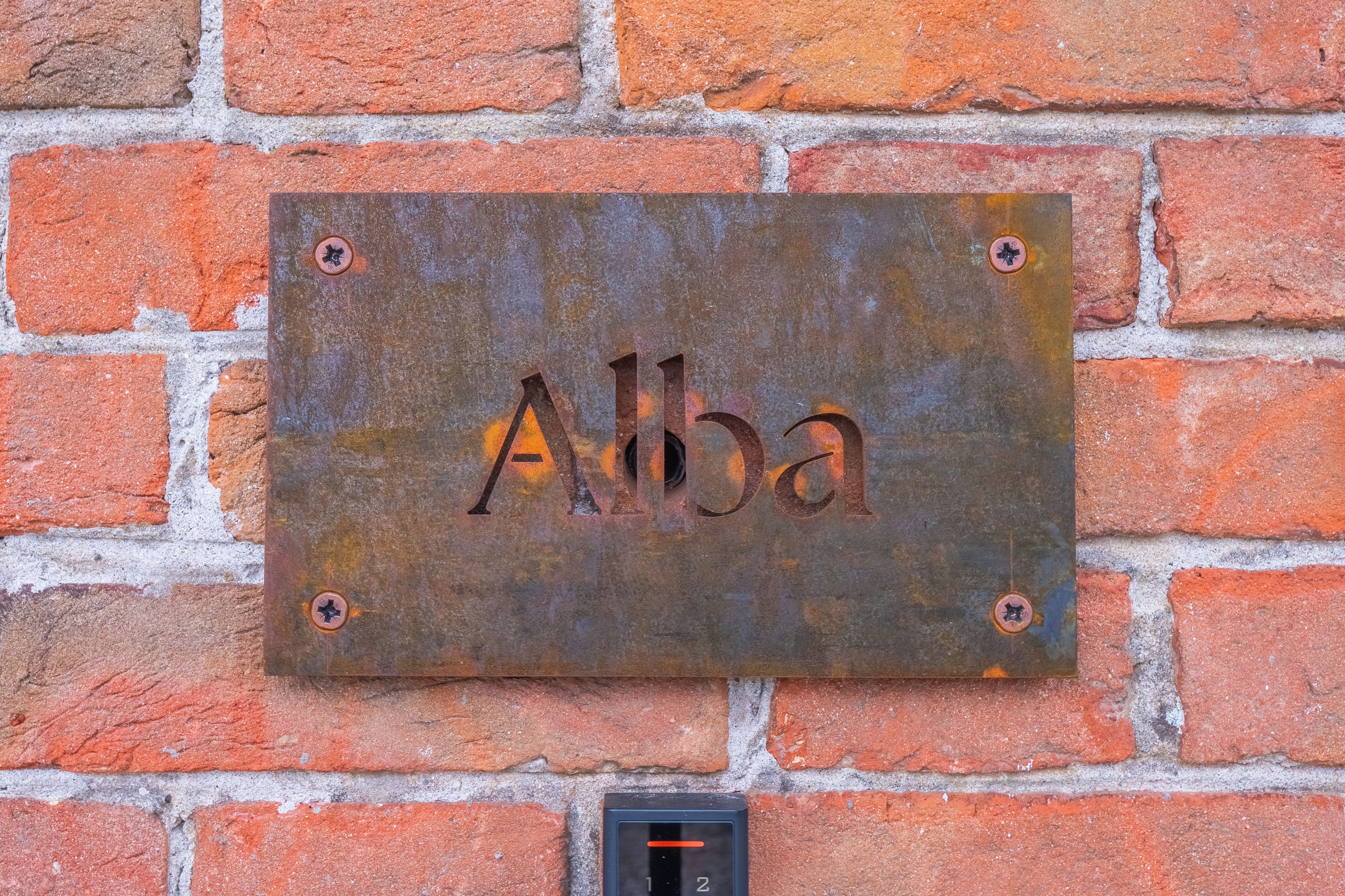Eine Metallplakette mit dem Schriftzug 'Alba' ist an einer Ziegelwand angebracht.