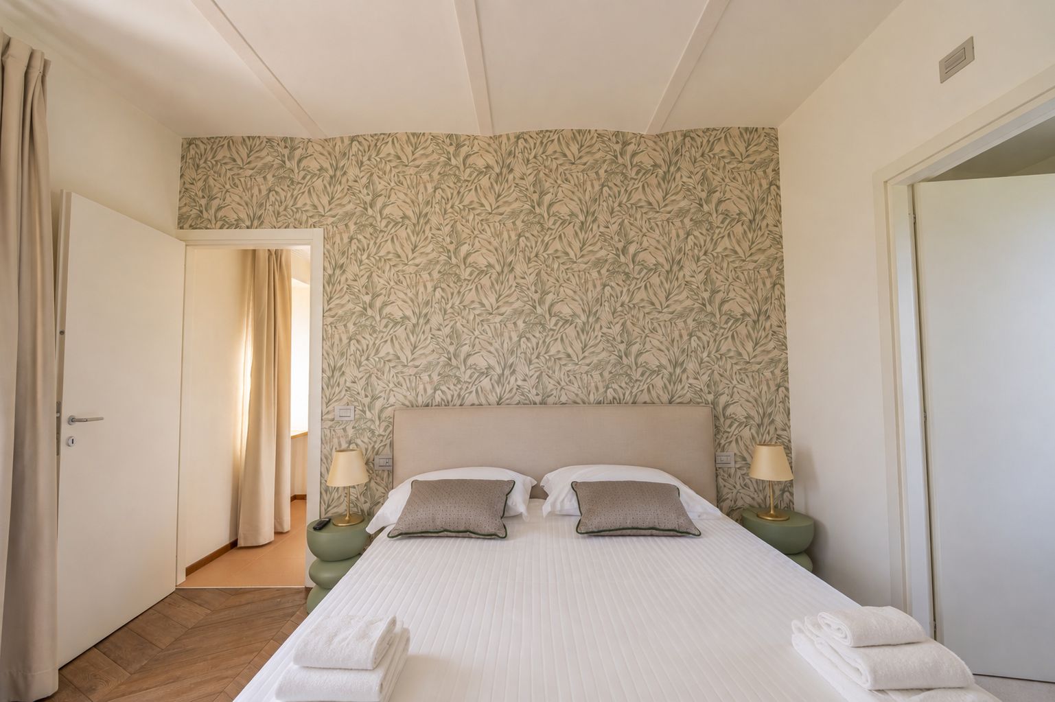 Una camera d'albergo con un letto grande, due comodini con lampade, una carta da parati decorata e una porta che dà su un'altra stanza.