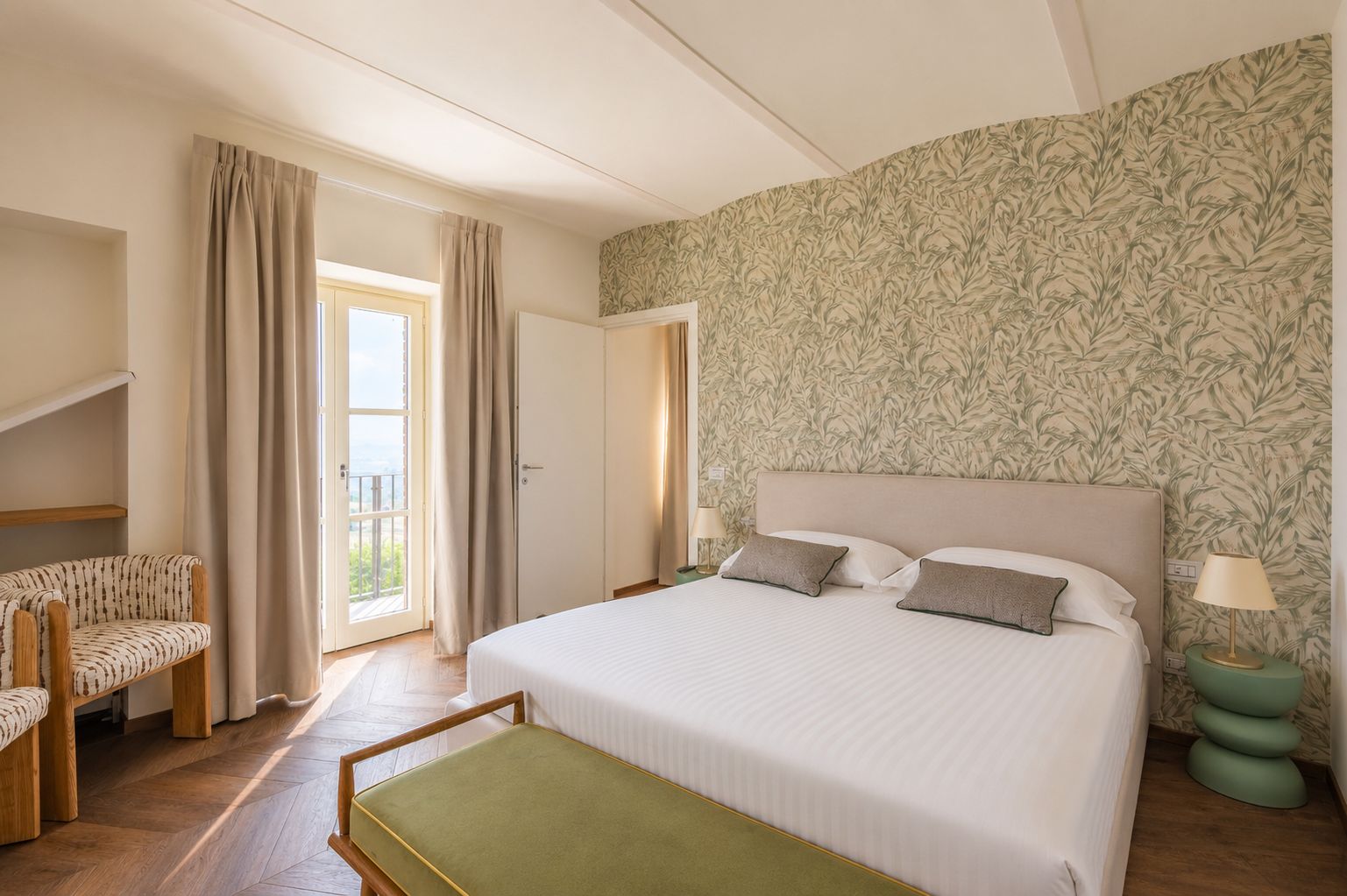 Quarto de hotel com cama, uma cadeira, uma mesa pequena, varanda e papel de parede estampado.