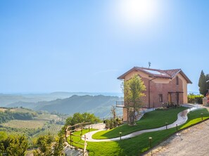 A house with a sloped roof, a brick exterior, and a paved driveway leading to a grassy area with a view of rolling hills.