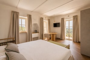 Quarto de hotel com cama, escrivaninha, cadeira, televisão e janelas amplas.