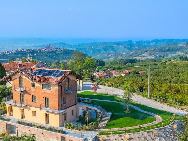 A modern two-story building with a brick facade and a solar panel roof, surrounded by a well-maintained lawn and a view of a hilly landscape.