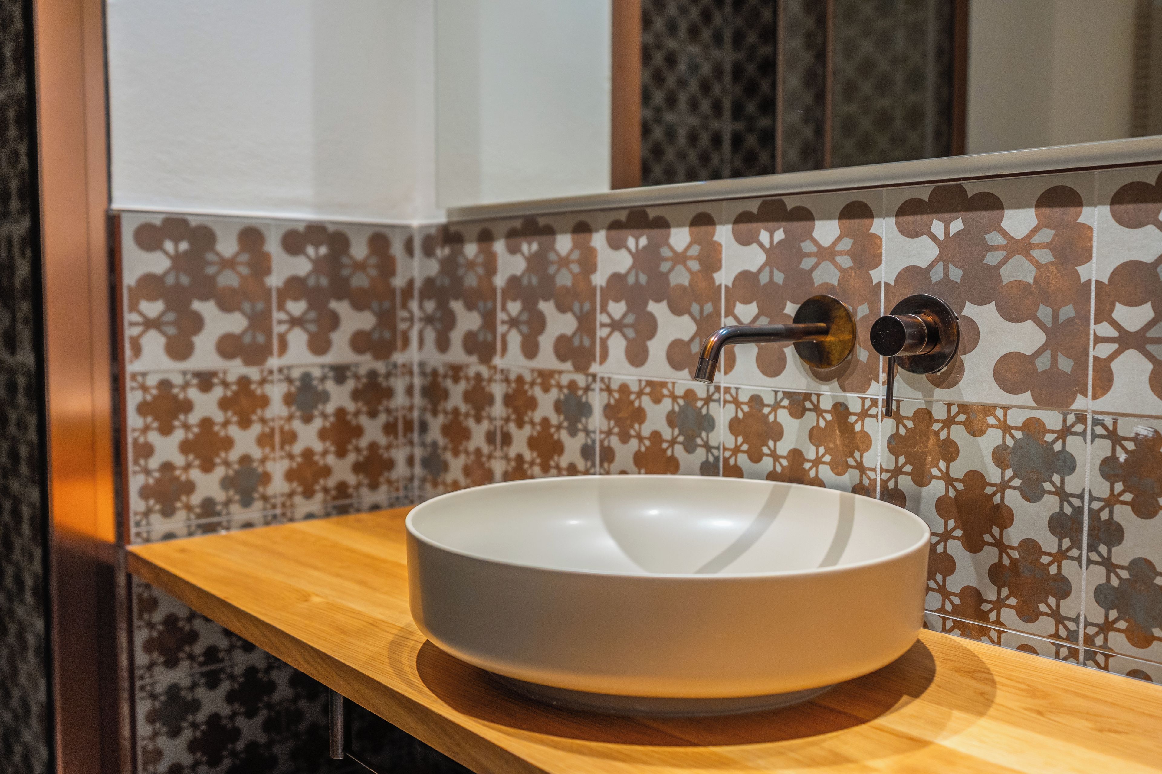 Un baño con un lavabo blanco sobre una encimera de madera, un grifo y baldosas estampadas en la pared.